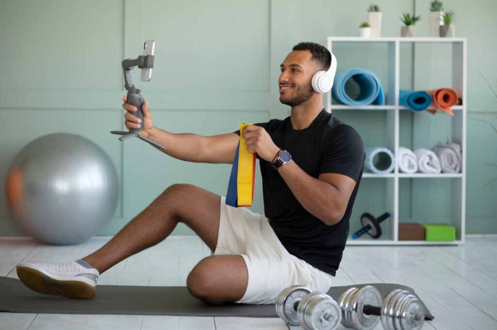 A man recording a fitness video using a smartphone stabilizer, wearing headphones, sitting on a yoga mat with fitness equipment like dumbbells and resistance bands.