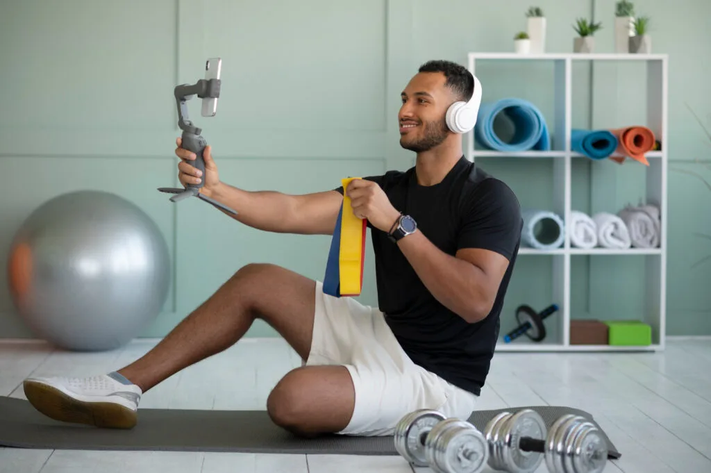 A man recording a fitness video using a smartphone stabilizer, wearing headphones, sitting on a yoga mat with fitness equipment like dumbbells and resistance bands.